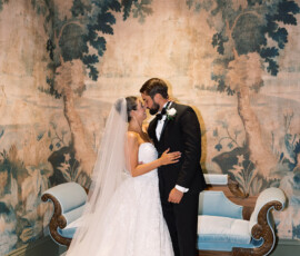 Bride and groom kiss in formal attire before an ornate loveseat, with a forest mural and elegant lighting at a New Orleans wedding.