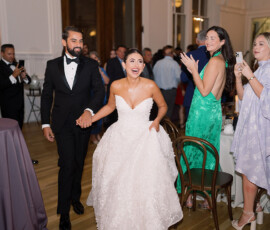 Bride and groom enter a New Orleans wedding reception, smiling and holding hands as happy guests clap and take photos.