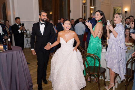 Bride and groom enter a New Orleans wedding reception, smiling and holding hands as happy guests clap and take photos.