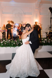 Bride and groom share a romantic first dance on a black and white floor with live band and floral arrangements at the reception.