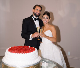 Bride and groom smile with champagne by a strawberry-topped wedding cake at their elegant New Orleans reception.