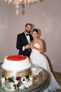Bride and groom smile with champagne by a strawberry-topped wedding cake at their elegant New Orleans reception.