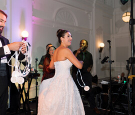 Bride in white gown smiles on stage with musicians; man in tux holds white beads, celebrating a lively New Orleans wedding.