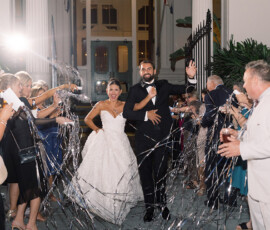 Bride and groom smile and wave among cheering guests with streamers at a lively New Orleans wedding at night.
