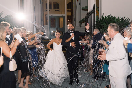 Bride and groom smile and wave among cheering guests with streamers at a lively New Orleans wedding at night.