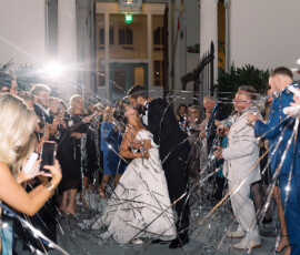 Bride and groom kiss outside at night as guests cheer, spray silver streamers, and capture the joyful Louisiana wedding moment.
