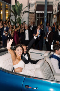 Bride and groom wave from blue convertible, cheered by guests outside CHOPHOUSE after classic Louisiana wedding.