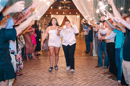 Newlywed brides making a joyful sparkler exit through cheering wedding guests inside a rustic barn venue at the end of the reception.