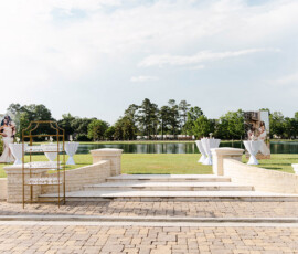 Lakeside Louisiana wedding setup with cocktail tables, stone steps, lawn, portraits, and scenic trees under a cloudy sky.
