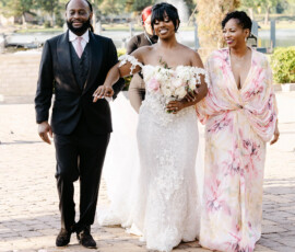 Bride in white gown with flowers walks outdoors, smiling with bridal party before elegant wedding ceremony on a sunny day.