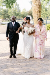 Bride walks outdoors with her parents while holding a bouquet of blush and white flowers before the ceremony.