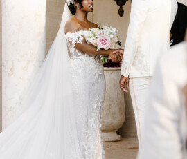 Bride in a white lace gown and groom in a white suit smile together at an outdoor Louisiana wedding ceremony.