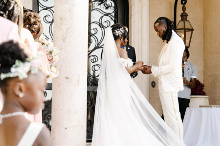 Bride and groom exchange rings during an outdoor wedding ceremony beside marble columns and flowing cathedral-length veil.