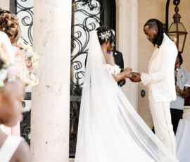Bride and groom exchange rings outdoors near ornate pillars, with bridesmaids, a flower girl, and classic Gulf South elegance.
