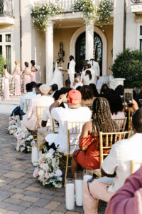 Guests watch as the bride and groom exchange vows during an outdoor ceremony beneath cascading floral arrangements at a grand estate entrance.