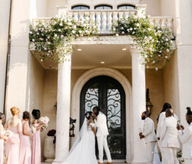 Bride and groom kiss on mansion steps with pink-clad bridesmaids, light-suited groomsmen, and floral-decorated columns.