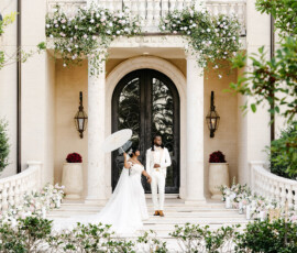 Bride with white umbrella and groom pose on floral mansion steps at their elegant New Orleans wedding, lush greenery and pool ahead.