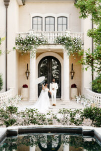 Bride and groom pose beside a reflecting pool outside a luxury estate decorated with lush blush and white floral installations.