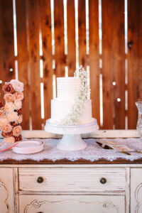 Simple white two-tier wedding cake decorated with delicate baby’s breath flowers displayed on a lace-covered table against a wooden barn wall.