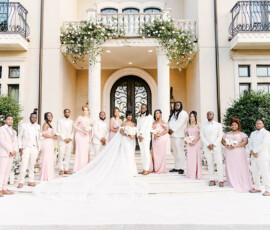 Bride, groom, and wedding party in pink and white pose on elegant mansion steps with white flowers and greenery in New Orleans.