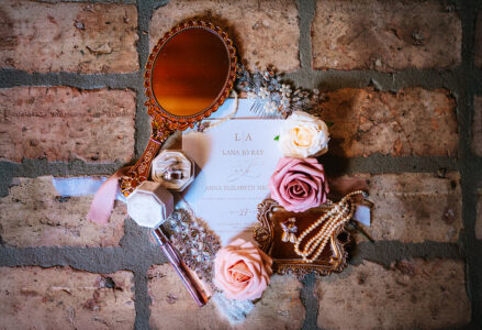 Bridal flat lay featuring wedding invitation suite, pearl jewelry, perfume, bridal hairpiece, pink roses, and rings arranged on a rustic brick floor.