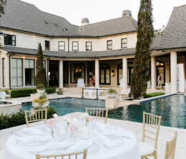 Chic outdoor poolside event at a large house with white-clothed tables, gold chairs, candles, and flowers on a sunny day.