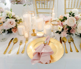 Gold utensils, pink napkin with brooch, white candles, and blush flower arrangements on a white table for a Louisiana wedding.