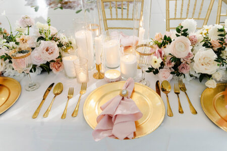Romantic wedding tablescape featuring blush roses, gold charger plates, pink napkins, candlelight, and gold flatware.