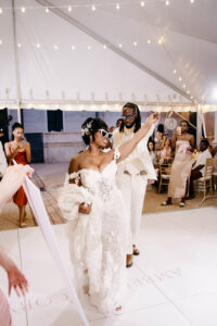 Bride in a lace off-the-shoulder wedding gown dances into the reception wearing white heart-shaped sunglasses beneath café lights.