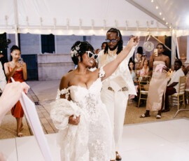 Bride in sunglasses and lace gown celebrates with drink as she and groom enter tented Gulf South wedding, guests and string lights above.