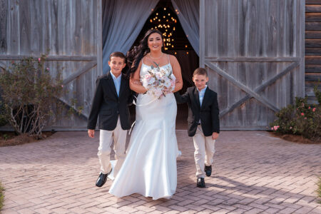 Bride walking hand-in-hand with two young boys in matching black jackets outside a barn venue while holding a blush and ivory bouquet.