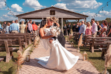 Newlywed brides sharing a kiss in the center aisle of their outdoor ceremony space beneath café lights and blue skies after exchanging vows.