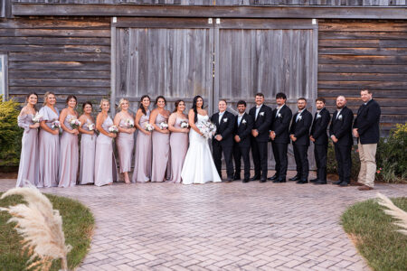 Full wedding party portrait in front of rustic wooden barn doors with bridesmaids in champagne satin dresses and groomsmen in black suits.