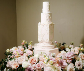 Elegant white wedding cake with floral decor on a round table amid pastel roses and greenery; perfect Louisiana wedding inspo.