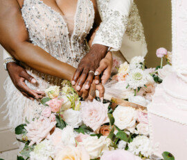 Smiling couple in elegant attire cuts pink wedding cake together, showcasing rings amid pastel flowers—New Orleans wedding inspiration.