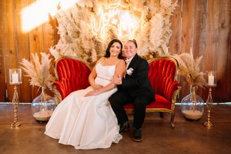 Newlywed brides seated together on a vintage red velvet loveseat beneath a glowing neon sign and pampas grass backdrop at their reception.