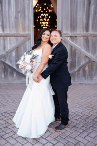 Two brides posing together in front of large wooden barn doors, one bride holding a pastel rose bouquet while the other embraces her from behind.