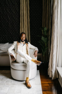 Groom in an ivory embellished tuxedo jacket sits confidently in a cream chair beside a sunlit window inside a luxury hotel suite.