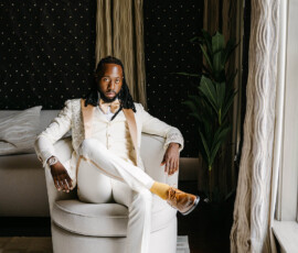 Man in a white suit with gold accents sits in a round chair, yellow socks and brown shoes, elegant Gulf South wedding scene.