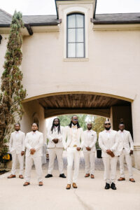 Groom and groomsmen pose beneath a grand covered entryway wearing coordinated ivory tuxedos with blush bow ties and brown dress shoes.
