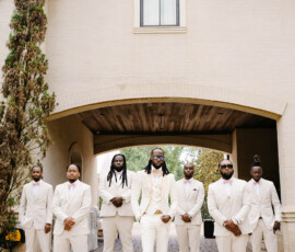 Seven men in white suits pose confidently in front of a light building with an archway, serious expressions, hands clasped.