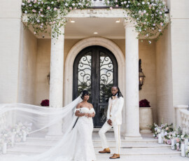 Bride and groom in white attire pose on mansion steps, flowers and columns around them, bride’s veil flowing to the side.