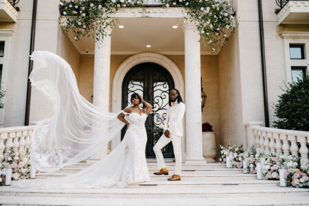 Bride’s cathedral veil billows dramatically beside the couple standing on the front steps of a floral-adorned mansion entrance.