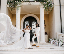 Bride in white gown and groom in cream suit pose on marble steps before ornate, flower-adorned New Orleans wedding venue.