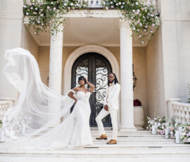 Bride and groom pose on mansion steps in New Orleans, her veil flowing amid flowers—timeless wedding inspiration.