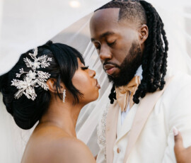Bride and groom under a white veil, gazing lovingly at their Gulf South wedding; she in off-shoulder dress, he in cream suit.