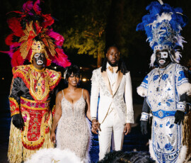 A couple in formalwear stands with two people in feathered costumes and face paint, holding umbrellas at a vibrant Louisiana wedding.