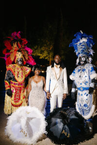 Bride and groom stand between two elaborately dressed Mardi Gras Indians during a nighttime wedding portrait in New Orleans.