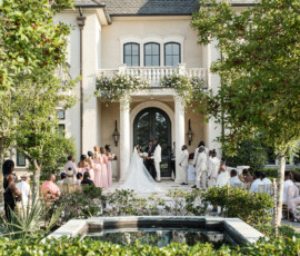 Bride and groom at an outdoor Louisiana wedding by a large house, with pastel-clad guests, greenery, and a small fountain.