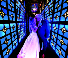 Bride and groom kiss beneath glowing chandeliers in a dramatic blue-lit corridor lined with stained-glass-inspired panels at their masquerade-themed wedding celebration.